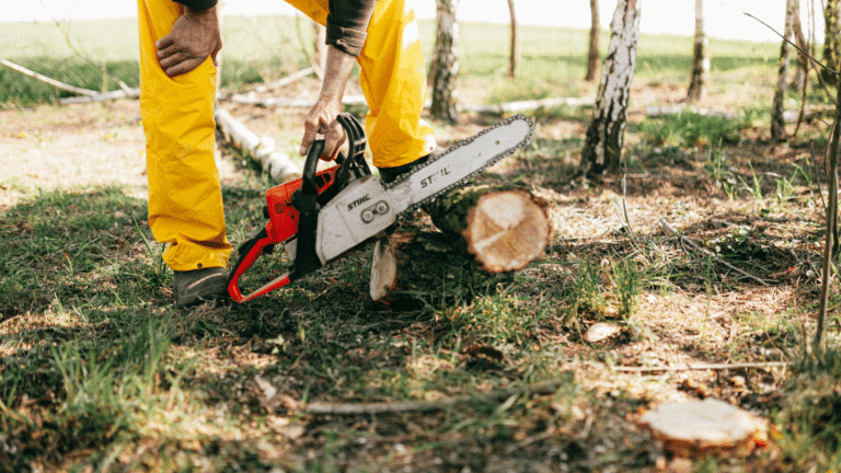 Person wearing yellow pants using a chainsaw to cut a log outdoors in a wooded area.